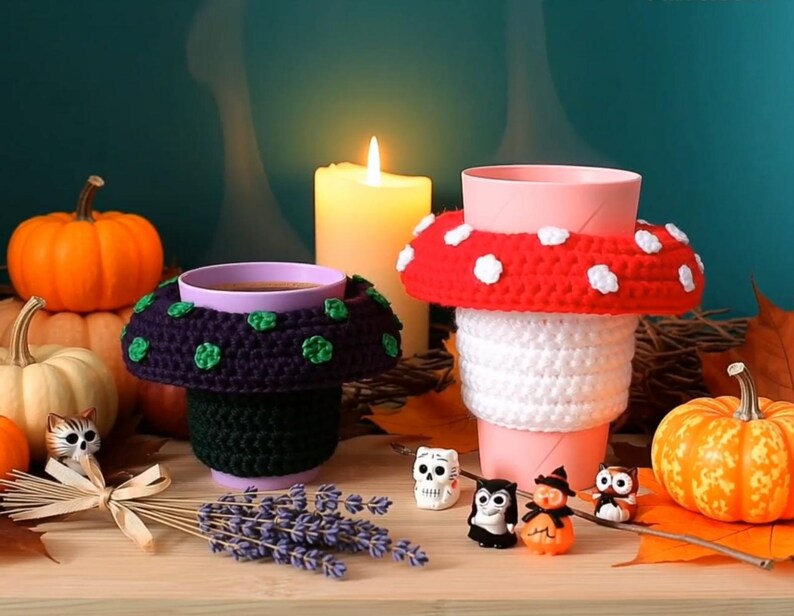 May include: Two crochet cup cozies shaped like mushrooms. One cozy is dark purple with green spots on a black base, and the other is red with white spots on a white base. They are placed around cups, surrounded by pumpkins, small figurines, and lavender.