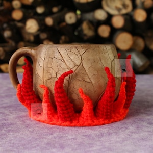 May include: A ceramic mug with a light brown, crackled glaze finish and a single handle, encased in a vibrant, crocheted cozy featuring a bright red and orange flame-like design that wraps around the base of the mug.
