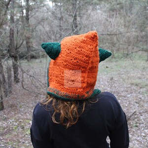 May include: A bright orange crochet hood featuring two dark green pointed ears on the sides. The hood is constructed with a textured stitch pattern and includes a dark green border along the bottom edge, covering the head and neck area.