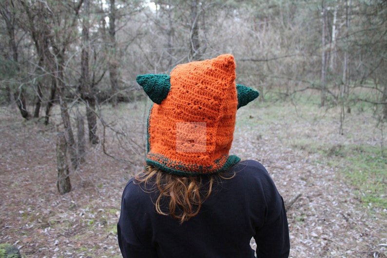 May include: A bright orange crochet hood featuring two dark green pointed ears on the sides. The hood is constructed with a textured stitch pattern and includes a dark green border along the bottom edge, covering the head and neck area.