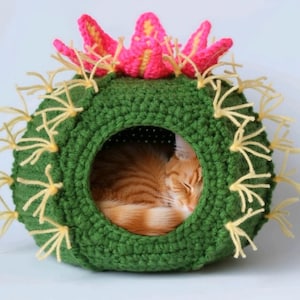 Puede incluir: Una cama para gatos hecha a mano con ganchillo en forma de cactus. La cama verde tiene espinas amarillas y una flor rosa y amarilla en la parte superior. Un gato naranja está durmiendo dentro de la cama.