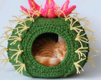 May include: A handmade crochet cat bed in the shape of a cactus. The green bed has yellow spines and a pink and yellow flower on top. An orange cat is sleeping inside the bed.