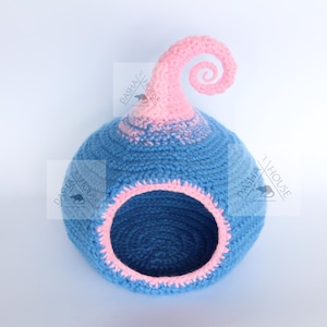 May include: A blue and pink crocheted pet bed with a whimsical design. The bed has a round opening and a decorative swirl at the top. The interior and opening are trimmed in pink.