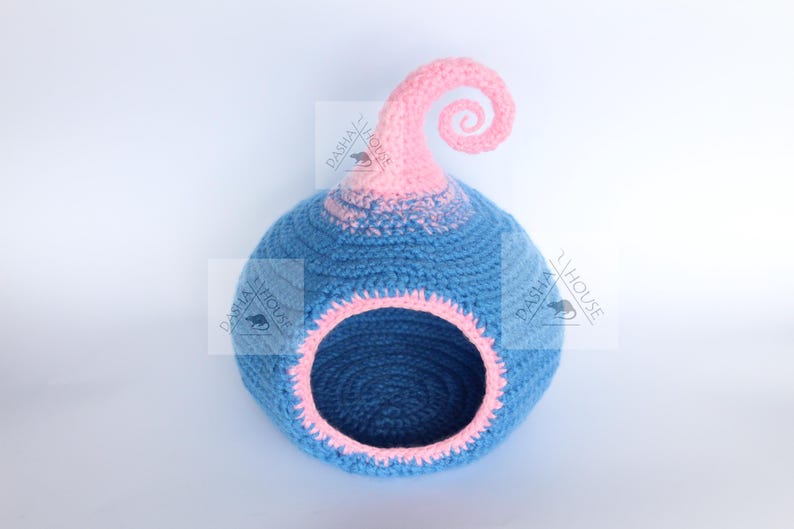 May include: A blue and pink crocheted pet bed with a whimsical design. The bed has a round opening and a decorative swirl at the top. The interior and opening are trimmed in pink.