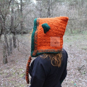 May include: A bright orange crochet hood with a dark green border and a small pointed ear on top. A long, braided orange and green tail hangs from the side. The word PATTERN is printed vertically in white letters on the left side of the image.