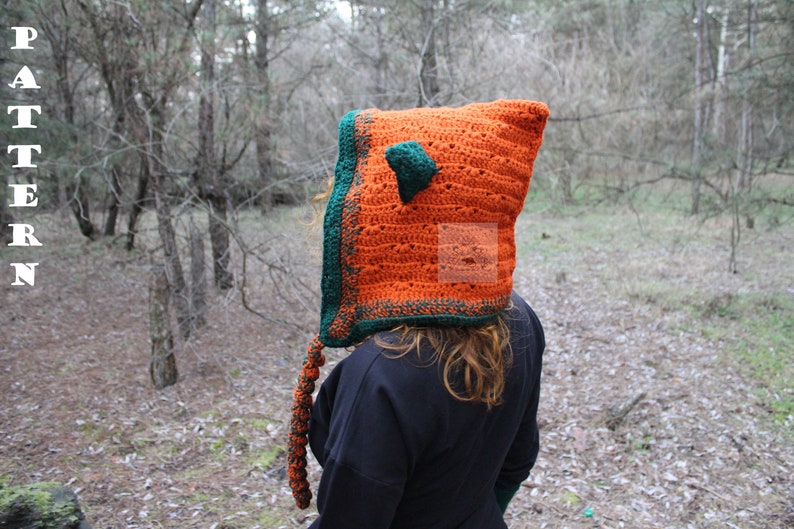 May include: A bright orange crochet hood with a dark green border and a small pointed ear on top. A long, braided orange and green tail hangs from the side. The word PATTERN is printed vertically in white letters on the left side of the image.