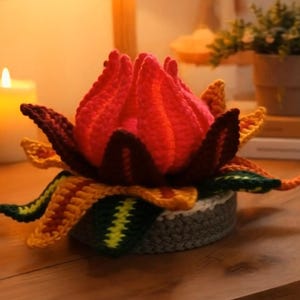 Pode incluir: Uma flor de crochê artesanal em tons de vermelho, rosa, amarelo, verde e marrom. A flor está colocada em um vaso cinza e branco. Uma vela acesa e uma pequena planta em vaso estão ao fundo.