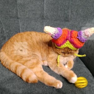 May include: An orange tabby cat wearing a colorful crocheted hat with horn-like projections. The hat features shades of purple, orange, green, and white. The cat is lying on a gray surface, with a yellow ball nearby. The word "PATTERN" is vertically displayed on the left.