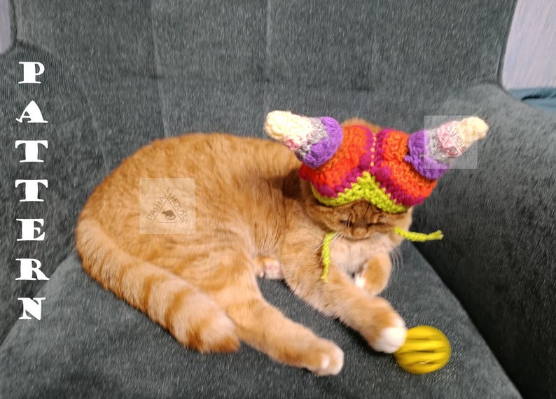 May include: An orange tabby cat wearing a colorful crocheted hat with horn-like projections. The hat features shades of purple, orange, green, and white. The cat is lying on a gray surface, with a yellow ball nearby. The word "PATTERN" is vertically displayed on the left.