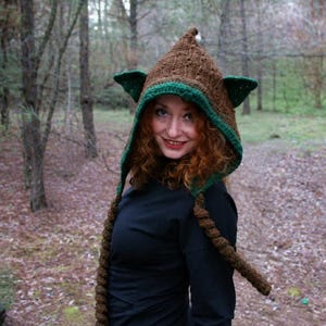 May include: A brown crocheted hood featuring a pointed top, two triangular green ears on the sides, and two long, thick braided tassels hanging down from the front. The hood has a green crocheted trim along the face opening.