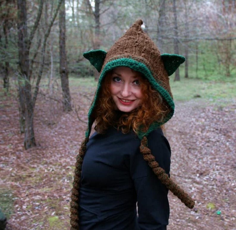 May include: A brown crocheted hood featuring a pointed top, two triangular green ears on the sides, and two long, thick braided tassels hanging down from the front. The hood has a green crocheted trim along the face opening.