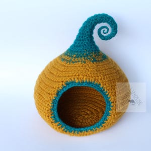 May include: A handmade crochet birdhouse in a teardrop shape. The birdhouse is primarily mustard yellow with teal blue accents around the opening and on the curled top. The textured design adds visual interest to this decorative item.
