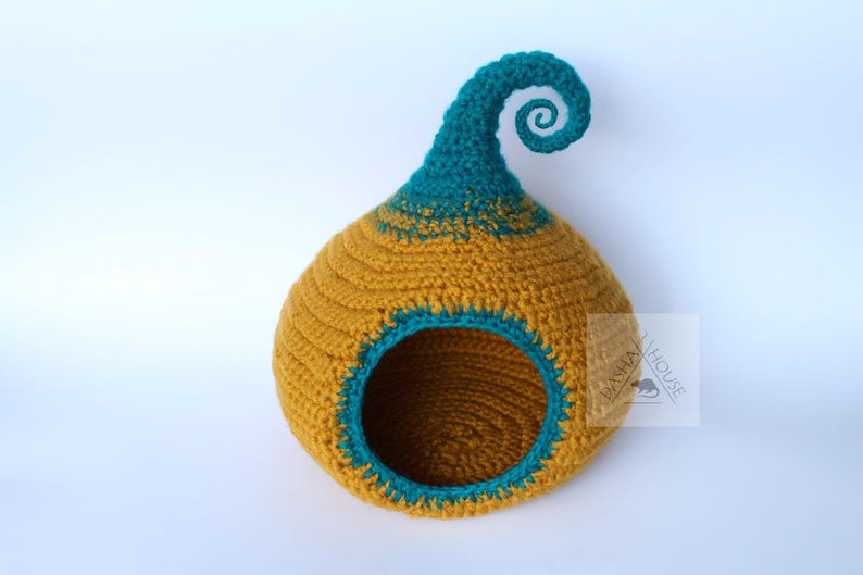 May include: A handmade crochet birdhouse in a teardrop shape. The birdhouse is primarily mustard yellow with teal blue accents around the opening and on the curled top. The textured design adds visual interest to this decorative item.