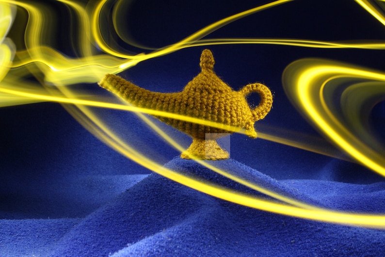 K&ouml;nnte beinhalten: Eine handgefertigte, goldgelbe H&auml;kellampe mit gebogenem Ausguss und Schlaufengriff. Die Lampe steht auf einer blauen Oberfl&auml;che, mit hellgelben Lichtstreifen, die von ihr ausgehen und einen magischen Effekt erzeugen.