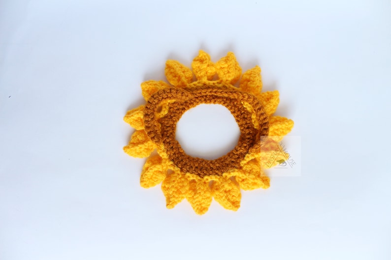 May include: A handmade crochet sunflower, featuring bright yellow petals and a brown center. The sunflower is designed to be worn as a headband or accessory. The craftwork is displayed on a white background.