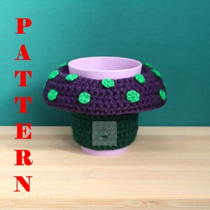 May include: A light purple cup featuring a crocheted mushroom-shaped cosy. The cosy has a dark green base and a wide, dark purple cap adorned with bright green polka dots. The word PATTERN is written vertically in red letters on the left side.