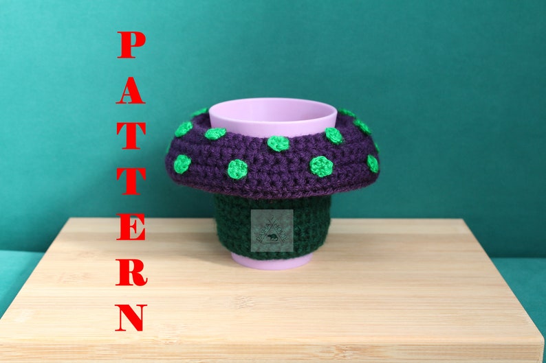 May include: A light purple cup featuring a crocheted mushroom-shaped cosy. The cosy has a dark green base and a wide, dark purple cap adorned with bright green polka dots. The word PATTERN is written vertically in red letters on the left side.