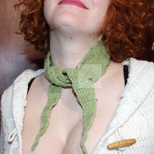 May include: A woman wearing a green knitted scarf around her neck. The scarf is tied in a loose knot at the front of her neck and the ends of the scarf hang down her chest.