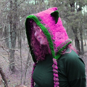 May include: A bright pink crochet hood featuring dark green trim, pointed cat-like ears, and two long braided tassels hanging from the front. The word PATTERN is printed vertically in pink on the left side of the image.