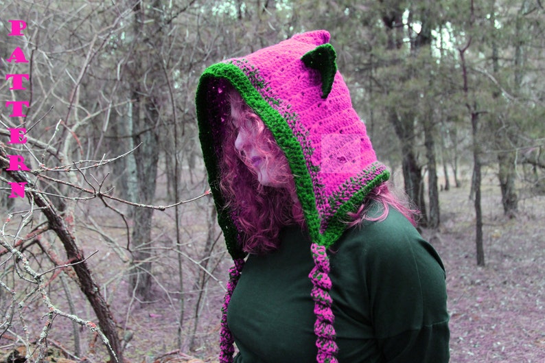 May include: A bright pink crochet hood featuring dark green trim, pointed cat-like ears, and two long braided tassels hanging from the front. The word PATTERN is printed vertically in pink on the left side of the image.