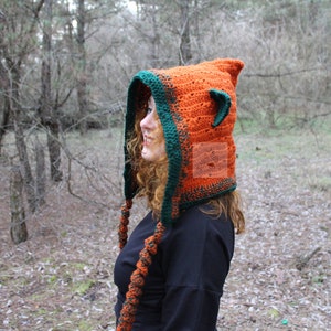 May include: A crochet hood in bright orange with a dark green border and small pointed ears on top. The hood features long, braided orange and green ties hanging down the sides. The word PATTERN is printed vertically in white on the left side.