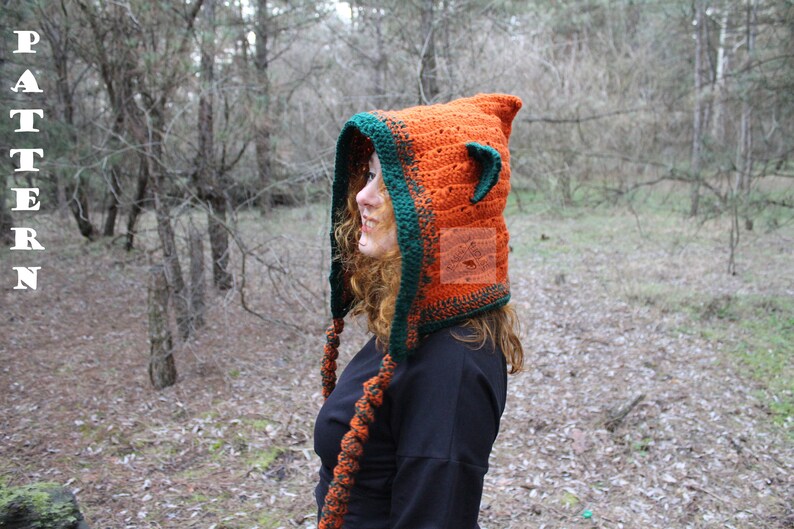 May include: A crochet hood in bright orange with a dark green border and small pointed ears on top. The hood features long, braided orange and green ties hanging down the sides. The word PATTERN is printed vertically in white on the left side.
