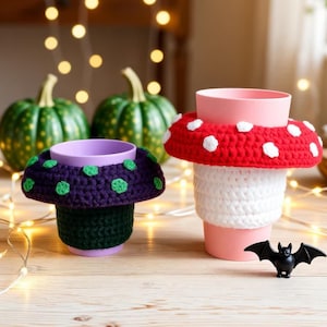 May include: Two plastic cups featuring crocheted mushroom-shaped covers. One cup has a dark purple base with green spots, while the other has a white base with a red cap and white spots. A small black plastic bat figurine sits in the foreground.