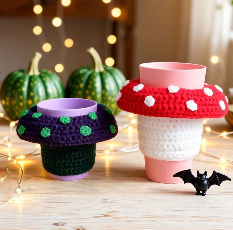 May include: Two plastic cups featuring crocheted mushroom-shaped covers. One cup has a dark purple base with green spots, while the other has a white base with a red cap and white spots. A small black plastic bat figurine sits in the foreground.