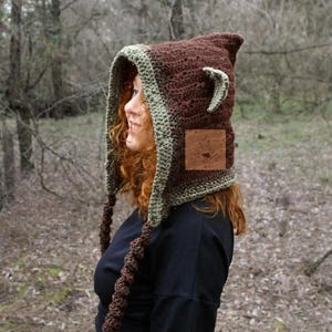May include: A brown crochet hood featuring a light green trim, pointed ears, and two long braided tassels hanging from the sides. A square brown leather patch with an embossed logo is attached to the side of the hood, which is worn over a black shirt.