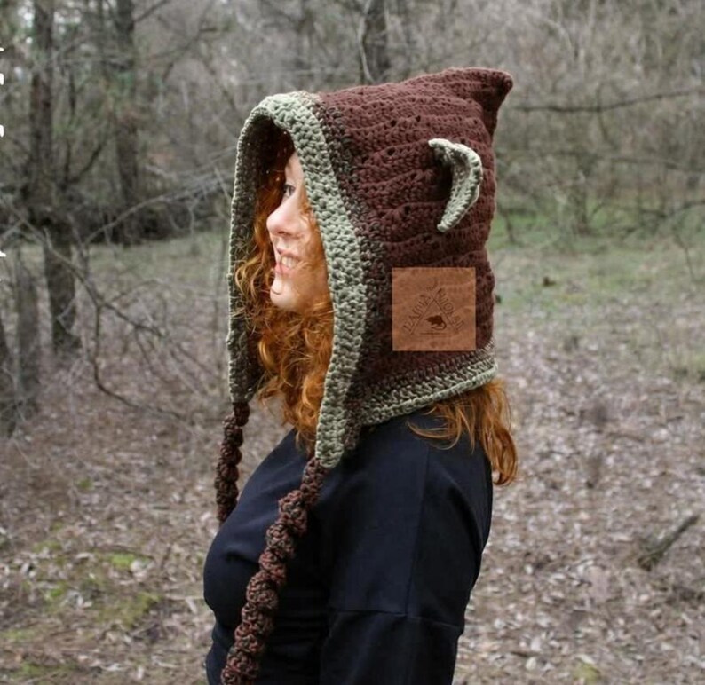 May include: A brown crochet hood featuring a light green trim, pointed ears, and two long braided tassels hanging from the sides. A square brown leather patch with an embossed logo is attached to the side of the hood, which is worn over a black shirt.
