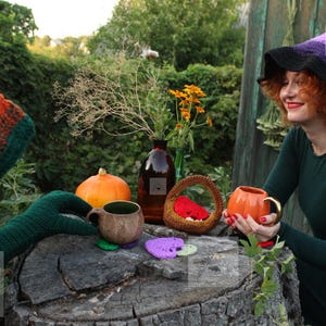 May include: A crocheted witch hat with a purple and black gradient, a dark green long-sleeved dress, a brown textured mug, a small orange pumpkin-shaped mug with a gold handle, a brown woven basket, and several small heart-shaped crochet coasters.