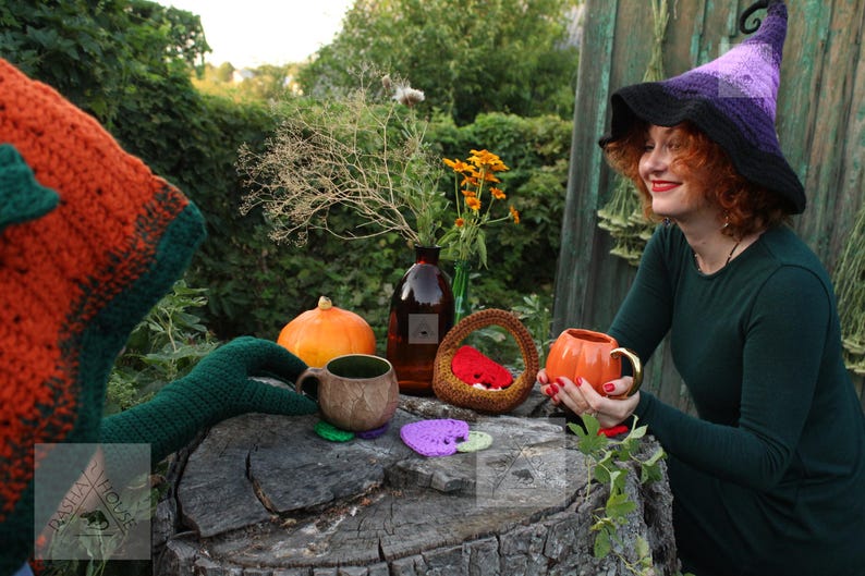 May include: A crocheted witch hat with a purple and black gradient, a dark green long-sleeved dress, a brown textured mug, a small orange pumpkin-shaped mug with a gold handle, a brown woven basket, and several small heart-shaped crochet coasters.