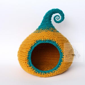 May include: A handmade crochet pet bed in a whimsical design. The bed is primarily mustard yellow with teal accents around the opening and on the curled top. The round opening provides easy access for a small pet.