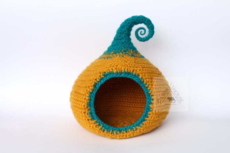 May include: A handmade crochet pet bed in a whimsical design. The bed is primarily mustard yellow with teal accents around the opening and on the curled top. The round opening provides easy access for a small pet.