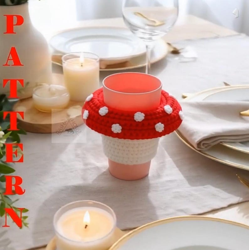 May include: A crochet cup cosy featuring a red mushroom cap design with white spots and a white stem base, wrapped around a light pink plastic cup. The word PATTERN is printed vertically in red on the left side of the image.