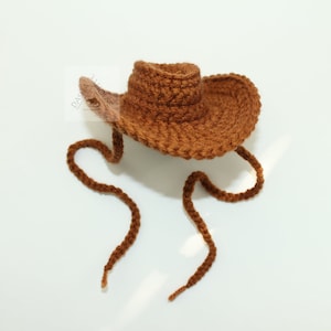 Puede incluir: Un sombrero de vaquero marr&oacute;n tejido a ganchillo, con una copa redonda y un ala ancha. El sombrero tiene una correa para la barbilla hecha con el mismo hilo. El sombrero est&aacute; sobre un fondo blanco.