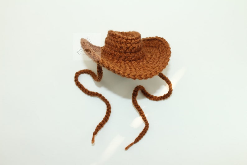 Puede incluir: Un sombrero de vaquero marr&oacute;n tejido a ganchillo, con una copa redonda y un ala ancha. El sombrero tiene una correa para la barbilla hecha con el mismo hilo. El sombrero est&aacute; sobre un fondo blanco.