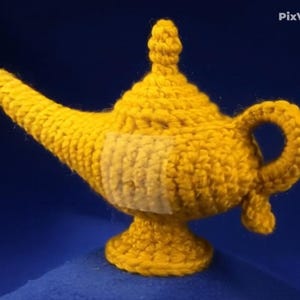 K&ouml;nnte beinhalten: Eine handgefertigte, gelbe H&auml;kellampe mit Ausguss, Griff und Sockel. Die Lampe hat ein strukturiertes, gewebtes Design und einen hellfarbigen Aufn&auml;her an der Seite. Der Hintergrund ist einfarbig dunkelblau.
