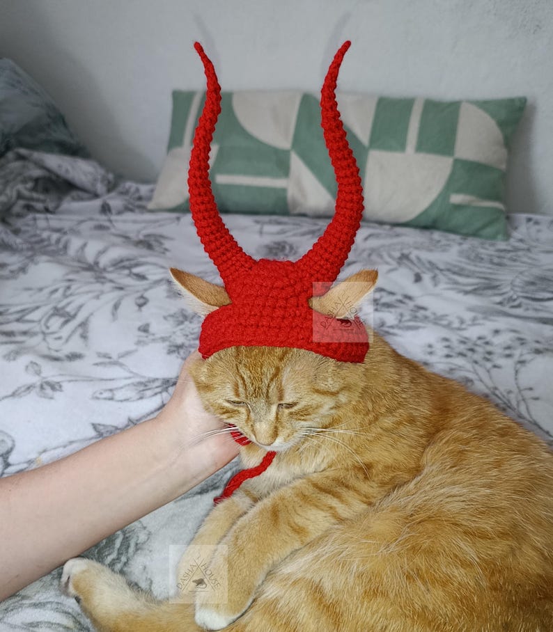 May include: A bright red crocheted hat designed for a cat, featuring two tall, curved, textured horns extending upward from the top. The hat includes ear holes and a thin red chin strap to secure it in place, crafted with a dense, uniform stitch pattern.