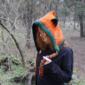 May include: A crocheted hood in bright orange with a dark green border and small pointed ears on top. The hood features long, braided orange and green yarn ties hanging from the front. The word PATTERN is printed vertically in red on the left side.