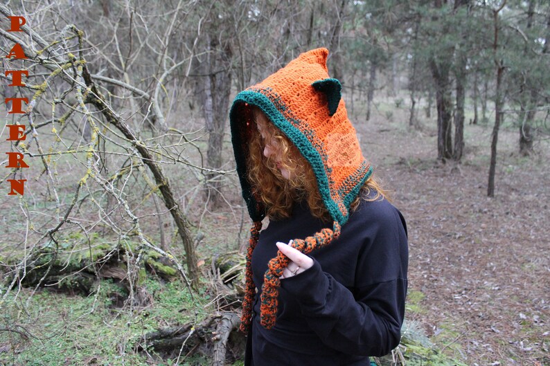 May include: A crocheted hood in bright orange with a dark green border and small pointed ears on top. The hood features long, braided orange and green yarn ties hanging from the front. The word PATTERN is printed vertically in red on the left side.