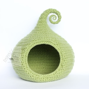 May include: A light green, crocheted pet bed shaped like a teardrop with a curled top. The bed has a circular opening and a small rectangular detail on the side. The texture of the crochet is visible.