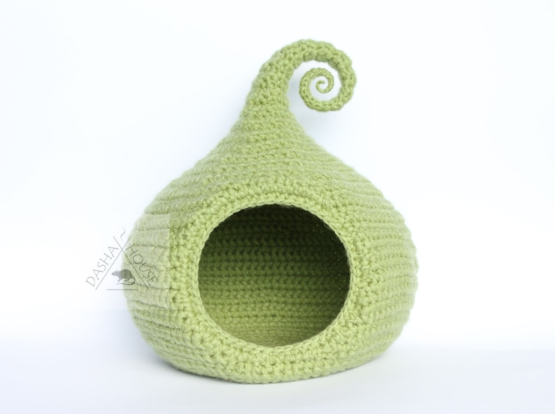 May include: A light green, crocheted pet bed shaped like a teardrop with a curled top. The bed has a circular opening and a small rectangular detail on the side. The texture of the crochet is visible.