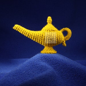 K&ouml;nnte beinhalten: Eine handgefertigte, gelbe H&auml;kellampe, die an eine Wunderlampe erinnert, steht auf einer blauen, strukturierten Oberfl&auml;che. Die Lampe hat einen gebogenen Ausguss, einen Griff und einen dekorativen Deckel. Das Wort "PATTERN" wird vertikal links angezeigt.