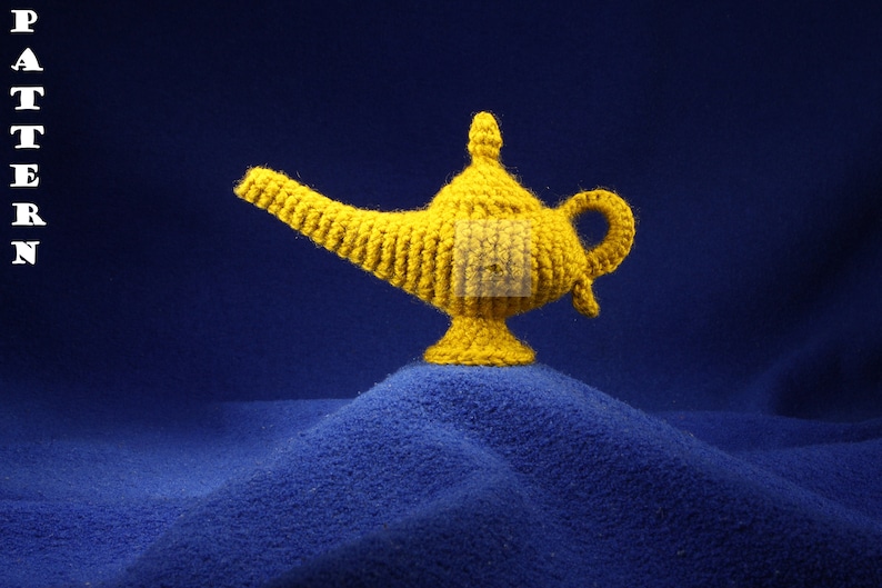 K&ouml;nnte beinhalten: Eine handgefertigte, gelbe H&auml;kellampe, die an eine Wunderlampe erinnert, steht auf einer blauen, strukturierten Oberfl&auml;che. Die Lampe hat einen gebogenen Ausguss, einen Griff und einen dekorativen Deckel. Das Wort "PATTERN" wird vertikal links angezeigt.