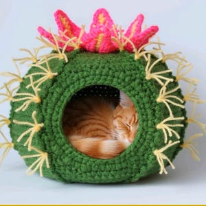 May include: A green crochet cat bed in the shape of a cactus, featuring yellow spines and a pink and yellow flower. An orange tabby cat is curled up asleep inside the bed. The bed is approximately 40 cm in diameter.