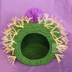 K&ouml;nnte beinhalten: Handgefertigtes H&auml;kel-Haustierbett in Kaktusform, gr&uuml;n mit gelben Fransen und einer lila Blume oben. Das Bett hat eine runde &Ouml;ffnung und steht vor einem lila Hintergrund. Ein einzigartiges Haustierzubeh&ouml;r.