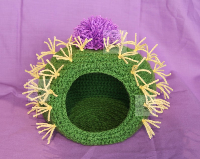 K&ouml;nnte beinhalten: Handgefertigtes H&auml;kel-Haustierbett in Kaktusform, gr&uuml;n mit gelben Fransen und einer lila Blume oben. Das Bett hat eine runde &Ouml;ffnung und steht vor einem lila Hintergrund. Ein einzigartiges Haustierzubeh&ouml;r.