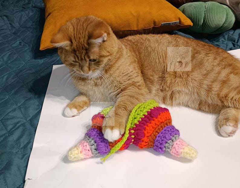 May include: A ginger cat with white paws interacts with a colorful, crocheted toy. The toy features a cone shape with a green top, transitioning to vibrant shades of pink, orange, purple, and yellow. The cat is lying on a white surface.