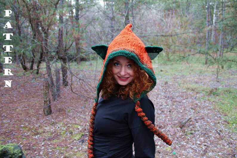May include: A crocheted hood with a pointed top and two triangular ear-like flaps, featuring a textured orange main section with a dark green border. Two long, braided orange and black tassels hang from the front. The word PATTERN is visible.